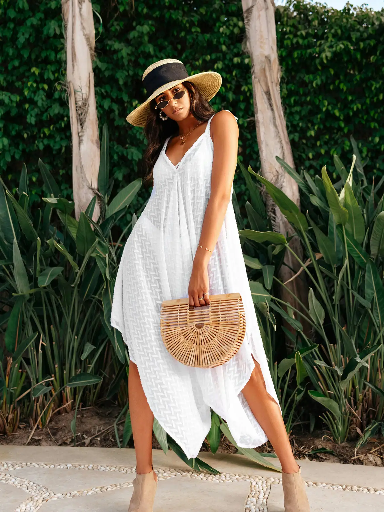 Woman in a white textured maxi dress and straw sun hat holding a bamboo bag beside tropical palms
