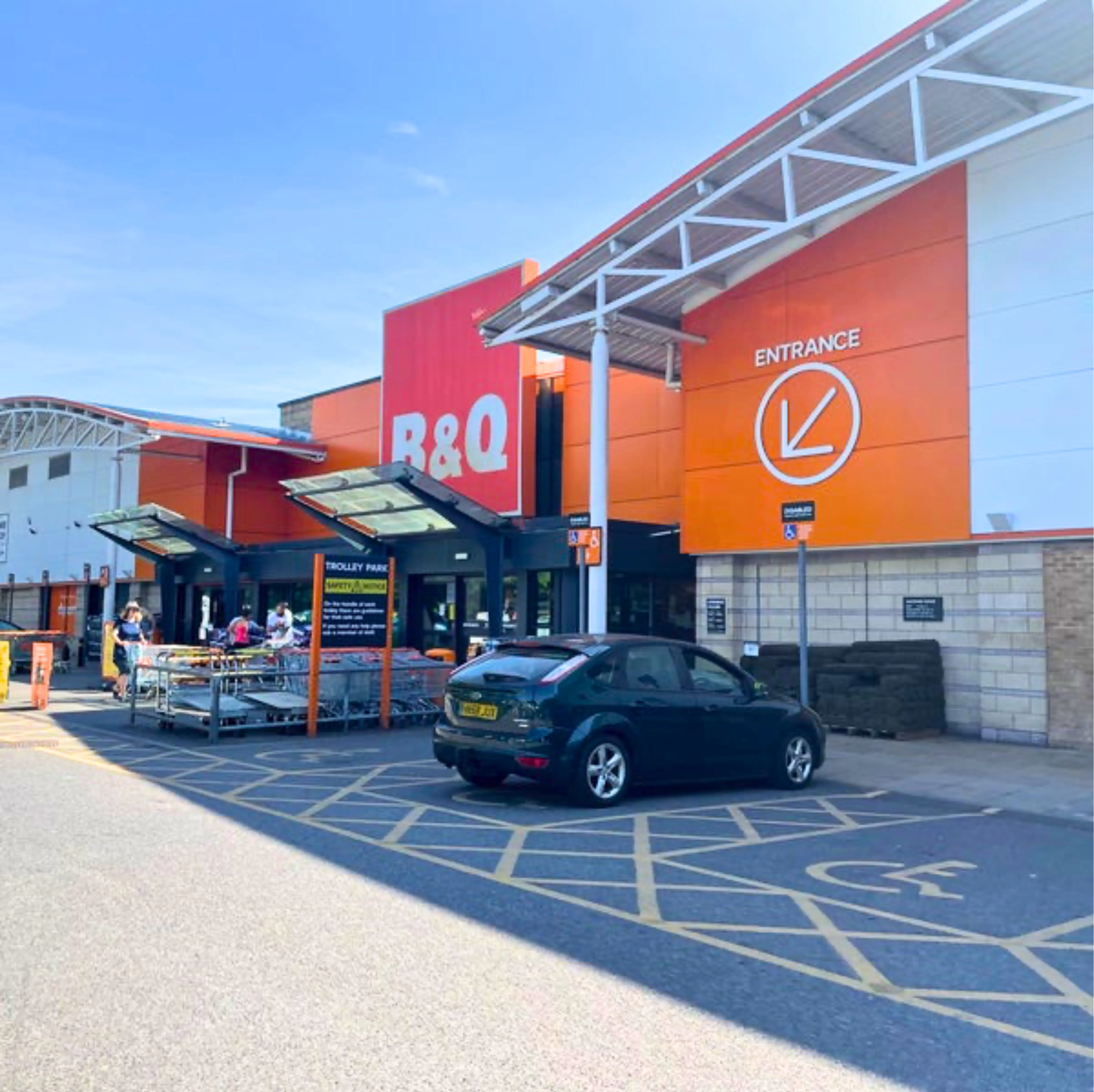 The image shows a large B&Q store under a clear blue sky, featuring a prominent orange and white exterior with an entrance sign, a parked dark green car, and shopping carts near the entrance.