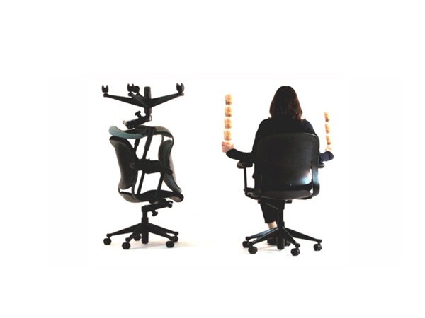 A woman sits in a black office chair with wheels, facing away from the camera. The chair is positioned against a white background. To the left, a black adjustable-height stand is folded and placed beside the chair. To the right of the chair, a black armrest is visible. The woman has long dark hair and is wearing a dark top and pants.