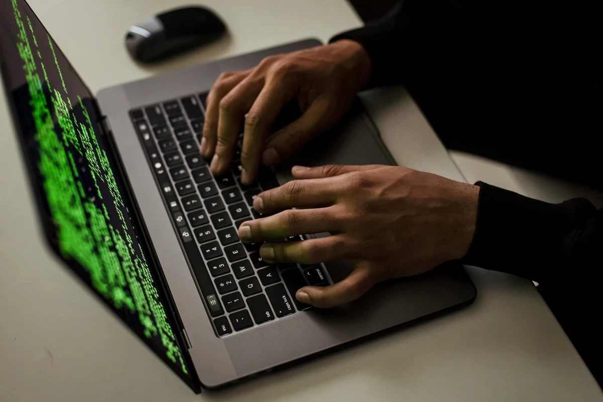Hands type on a laptop keyboard displaying green code on the screen. A computer mouse sits beside the laptop on a clean desk.