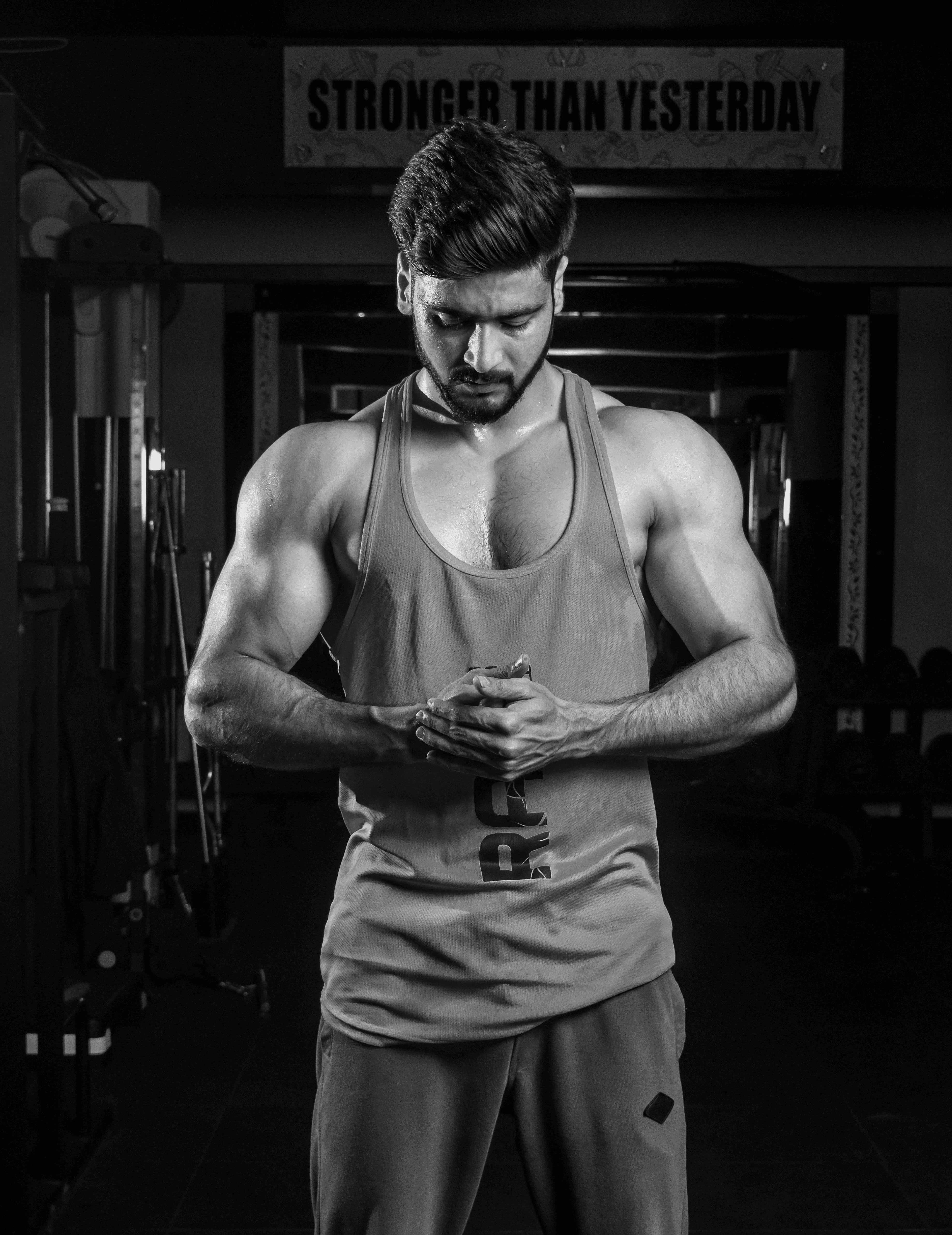 A muscular man in a gym wearing a sleeveless tank top stands with hands clasped, showcasing his defined arms and focused expression, with gym equipment and a motivational banner reading "Stronger Than Yesterday" in the background.