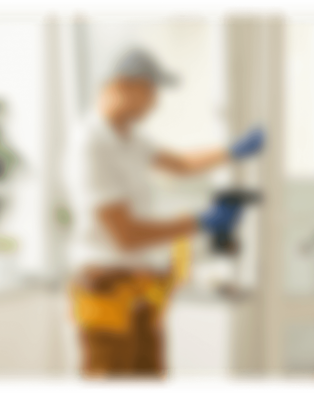 Blurred photo of a worker in a cap and tool belt installing a door hinge.