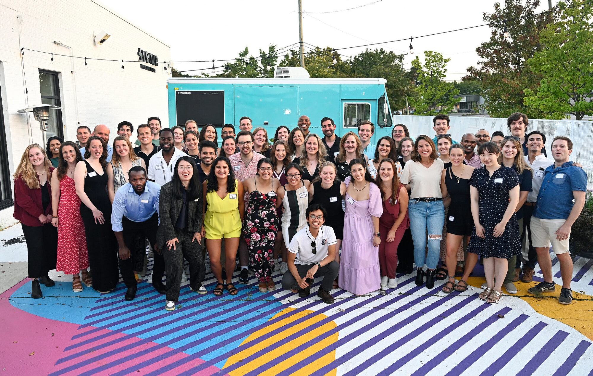 A large group of diverse people posing together outdoors on a colorful patterned surface.