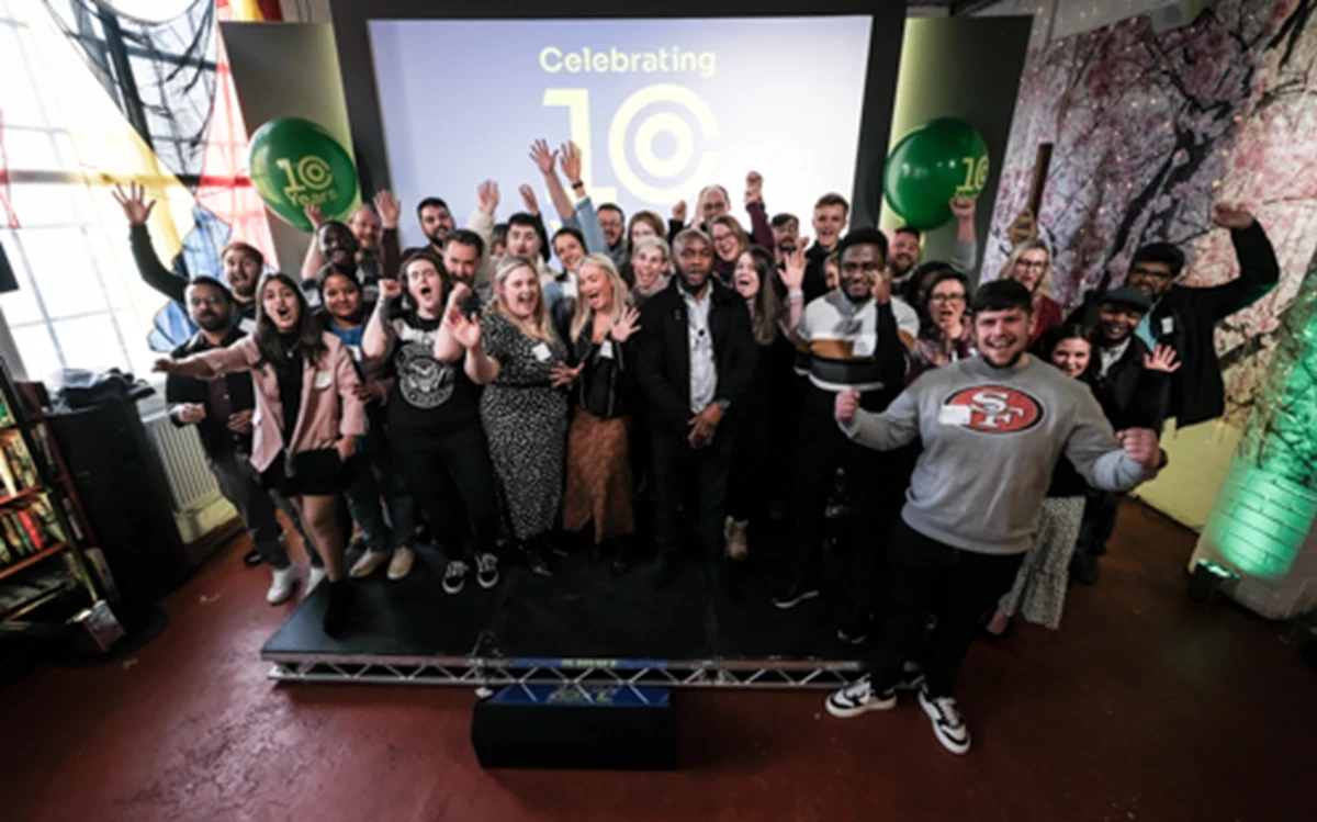 A large group of people standing on a small stage in an indoor venue, posing together with raised hands in a celebratory gesture. Behind them is a large screen displaying the words “Celebrating 10” with a circular design, and two green balloons on either side with the number “10” printed on them. The setting includes colorful wall art on the right and a bookshelf on the left, with bright natural light coming through a window. The floor is red, and the overall atmosphere suggests a festive event marking a 10-year anniversary.