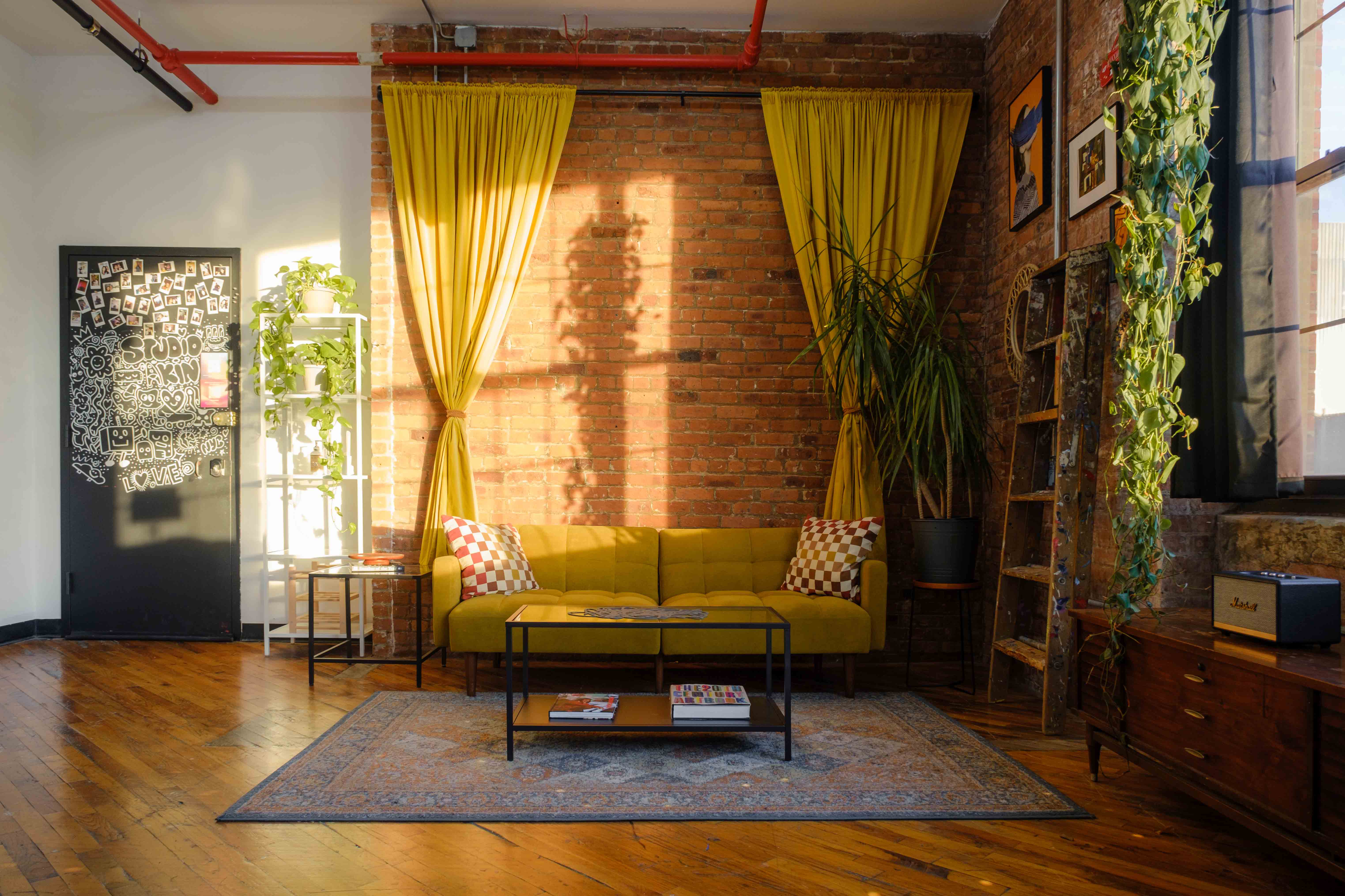 A sunlit room with exposed brick walls features a yellow couch with checkered pillows, vibrant plants, and eclectic decor, creating a cozy and inviting atmosphere. Natural light streams in from a large window with yellow curtains, adding warmth to the space.