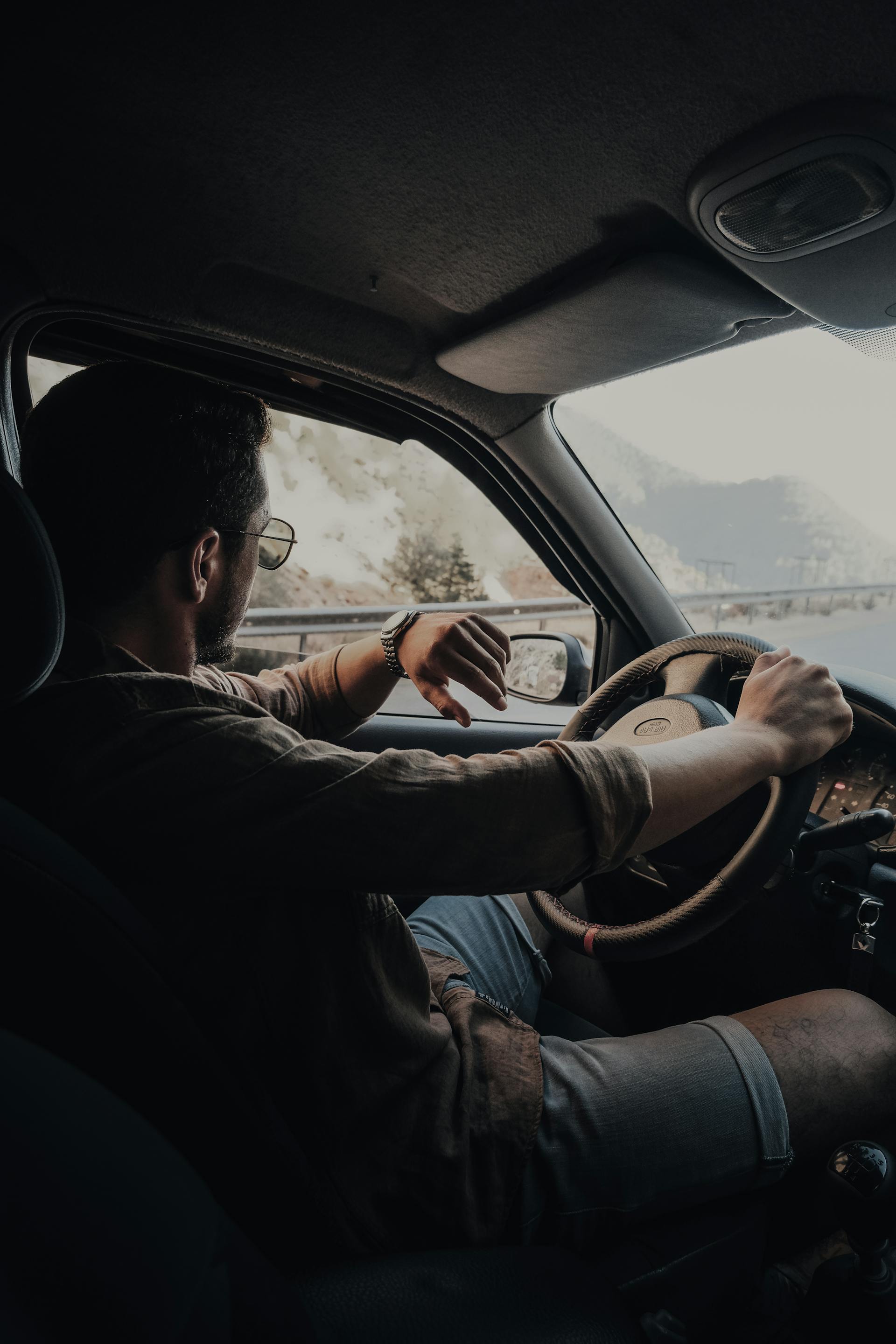 A man with sunglasses and a wristwatch is driving a car on a road, seen from the passenger's seat. He is wearing a button-down shirt and shorts. His right elbow is resting on the open window frame, and both hands are on the steering wheel. Hilly, mountainous terrain is visible outside the window.