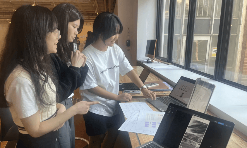 Three students gathered at a window-side desk reviewing papers and laptops during a design session.