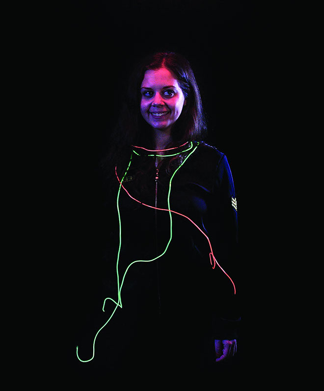 A woman with long brown hair stands against a black background. She wears a black zip-up jacket with a neon green and red neon cord draped around her neck and shoulders. The cord trails down her chest and ends near her waist. The woman’s face is illuminated with pink and purple neon light, and she has a slight smile. She is looking directly at the camera.