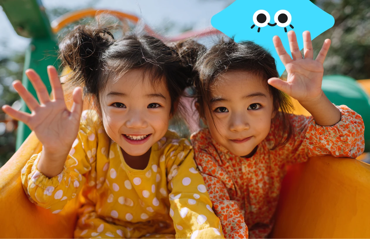 Two smiling children in polka dot outfits wave while sitting on a colorful slide on a sunny day. A playful blue cartoon face is in the top corner.