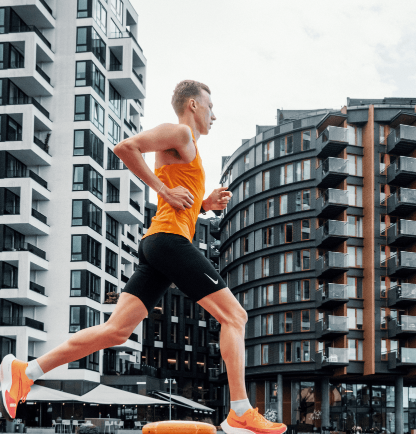 A runner wearing an orange singlet and black shorts and orange sneakers running through a city landscape with tall buildings in the background. 