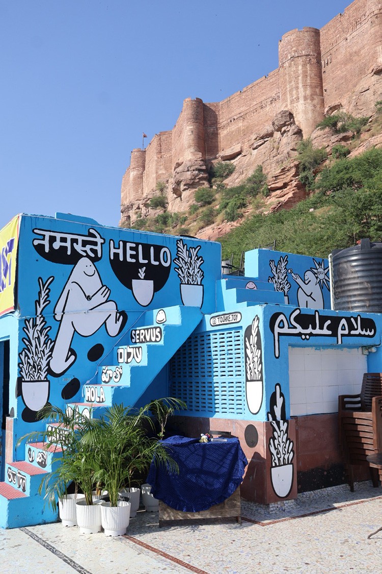 Mural painting with joyful cartoon characters and the word ‘Hello, नमस्ते, سلام عليكم’ painted on a terrace of a blue building near Jodhpur Fort.