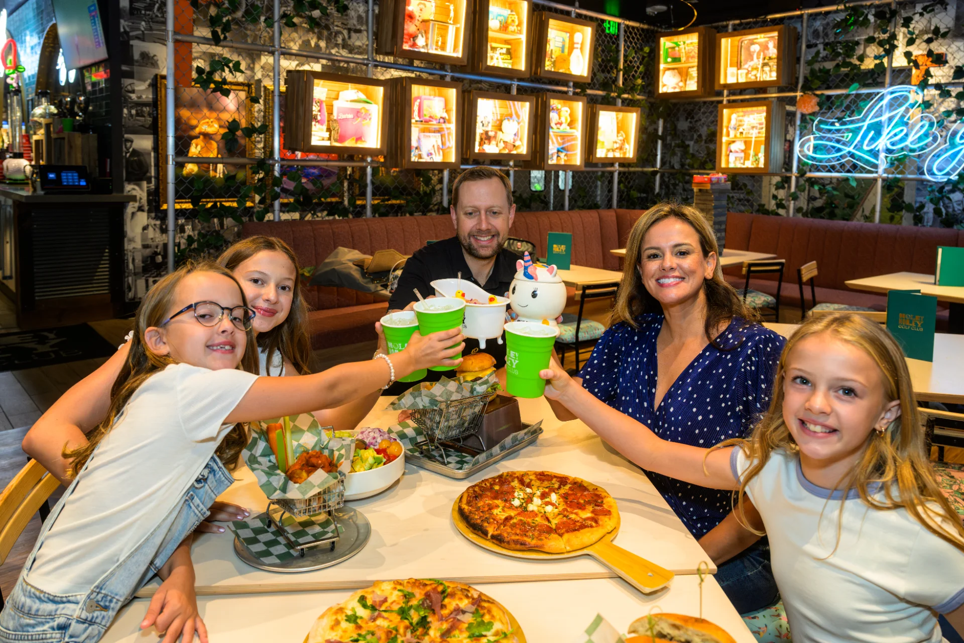 Family clinking cups around a table of pizza and salad in the neon-lit Holey Moley dining room