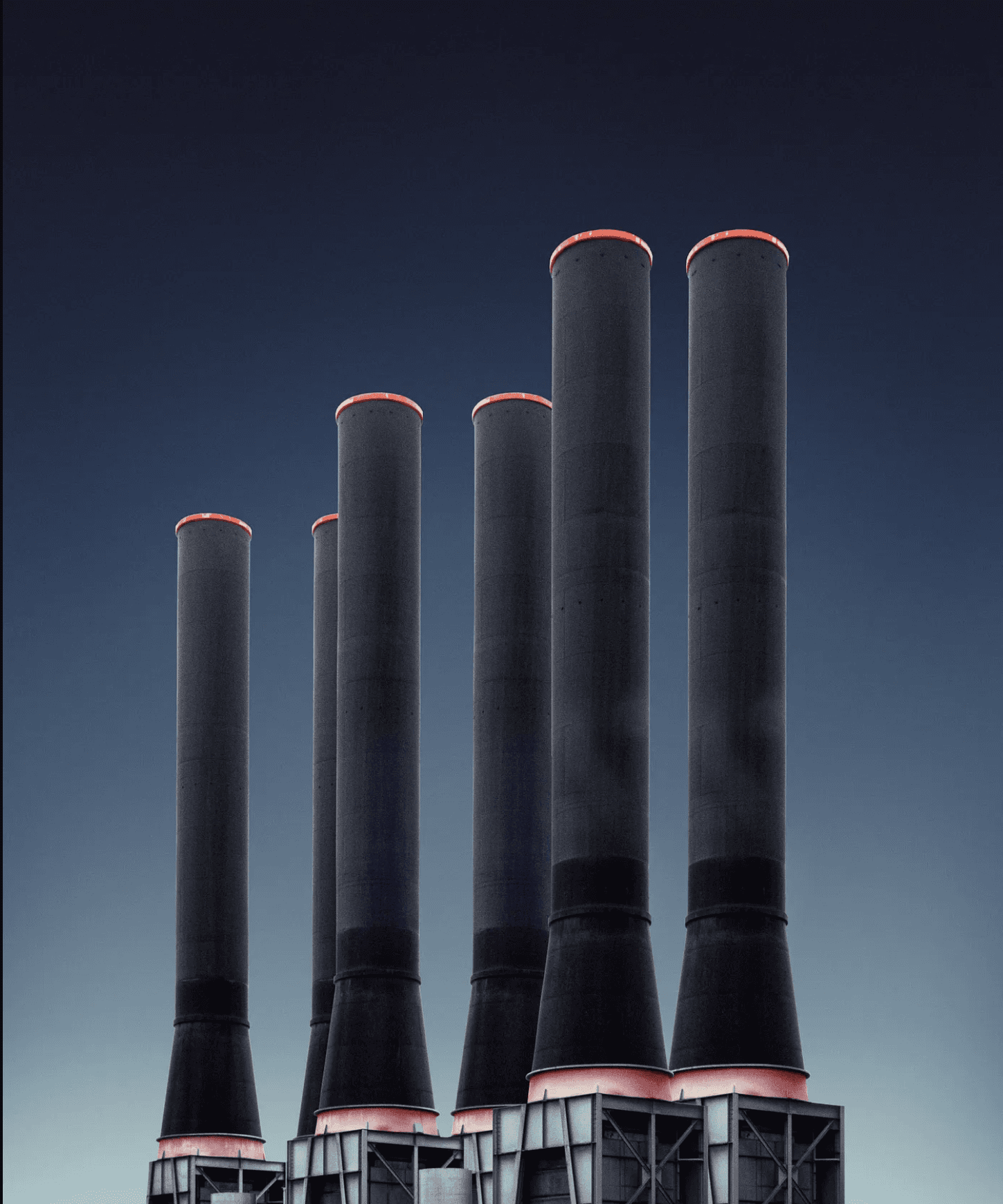 Photograph of a power plant's multiple tall, cylindrical smokestacks against a clear blue sky, with red warning lights on the stacks' rims