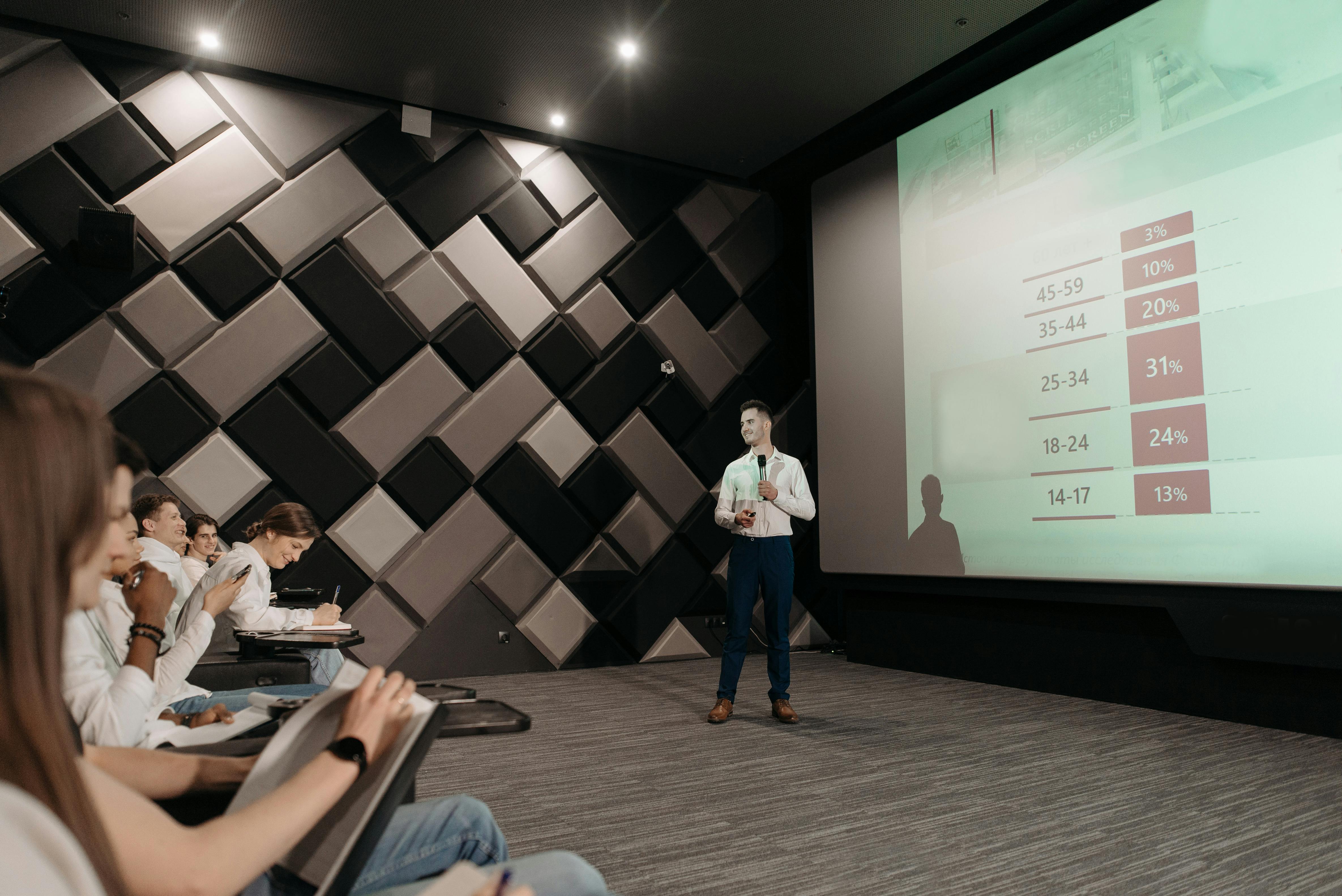 Relatore che illustra un grafico su un grande schermo durante una presentazione, mentre il pubblico prende appunti.