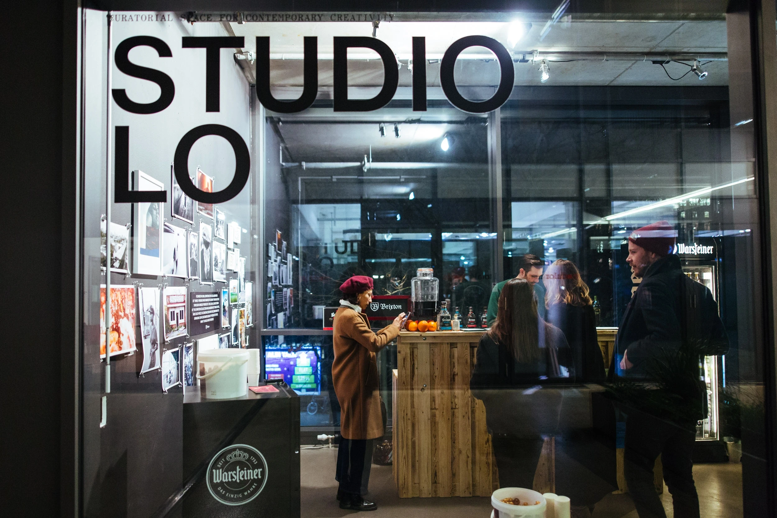 View through the STUDIO LO window lettering into the BRIXTON pop-up at night, with guests at the wooden bar and a photo wall.