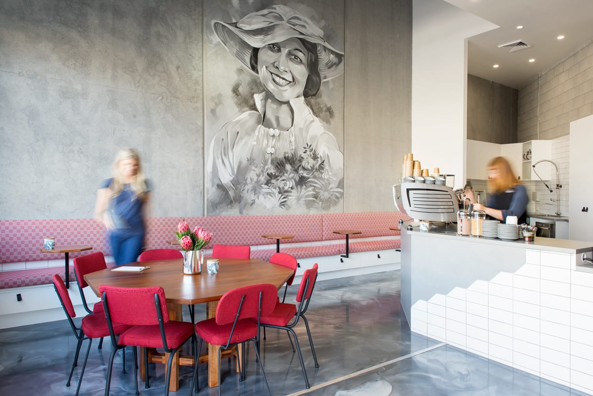 Contemporary cafe interior design with a large portrait mural, red upholstered dining chairs, long banquette seating, and a white-tiled coffee counter.