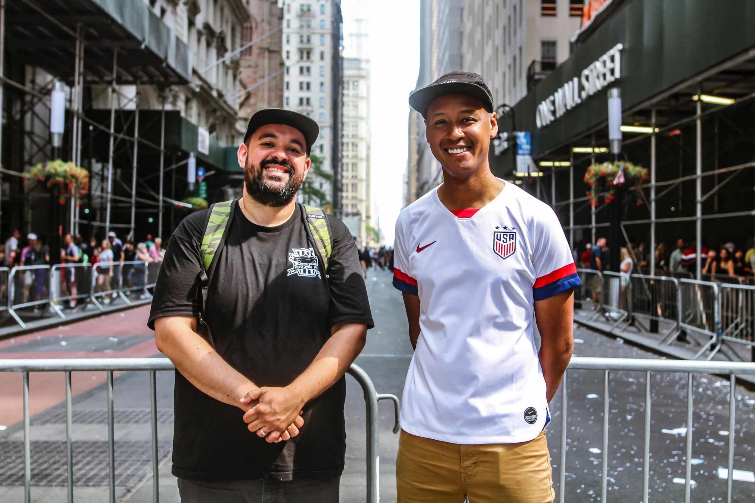 The Cooligans stand smiling on a city street, with metal barricades and crowds in the background. One wears a black shirt and cap; the other wears a white USA soccer jersey and black cap. Tall buildings and scaffolding line the street.