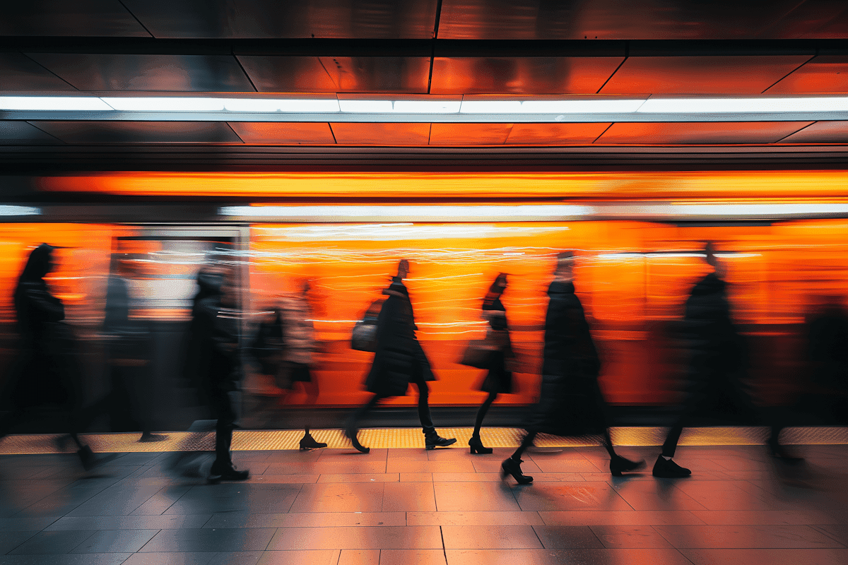 A diverse group of people walking through a busy subway station, with signs and trains visible in the background.