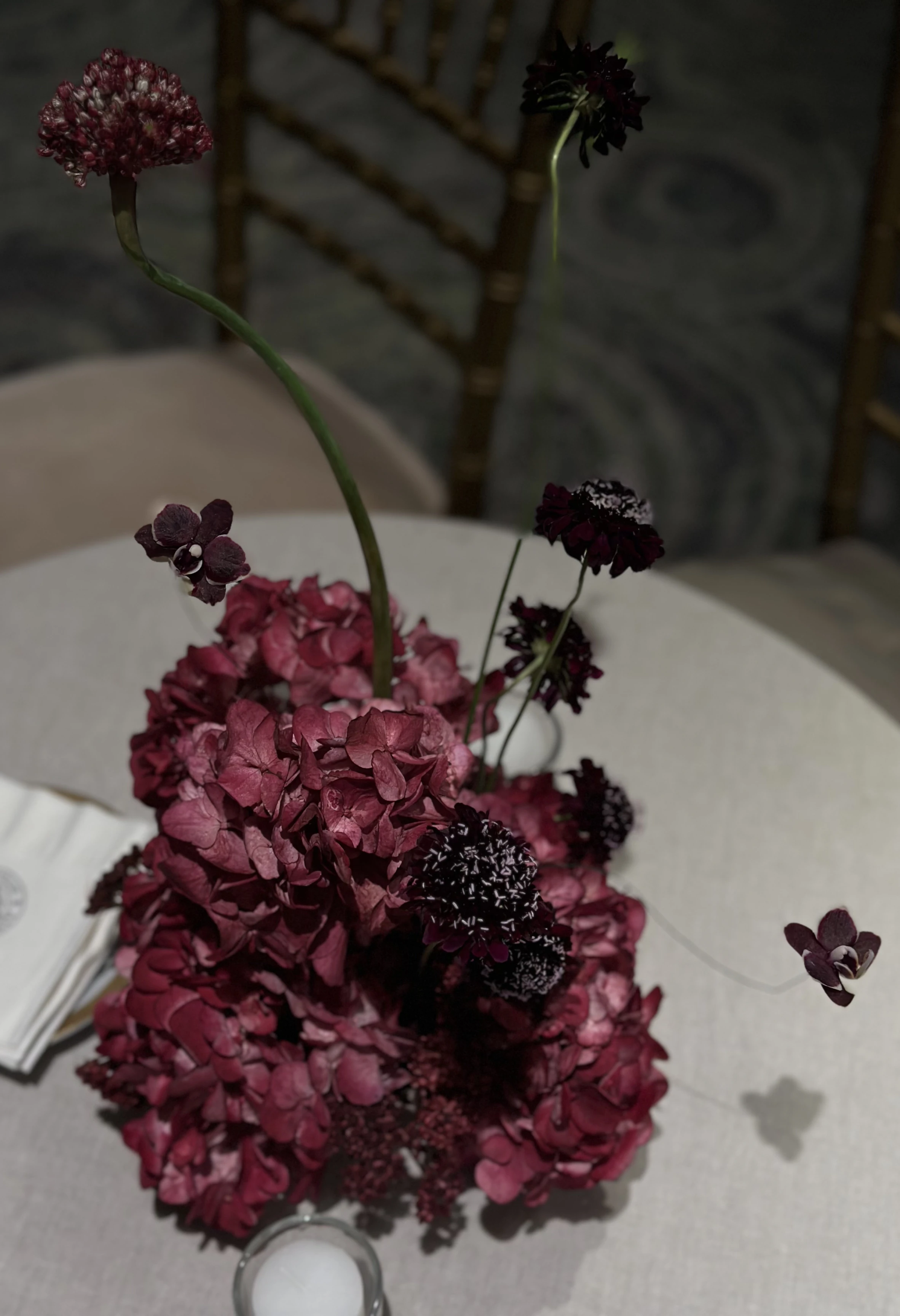 Vista cenital de arreglo floral inmersivo con hortensias color vino y orquídeas sobre mantel de lino gris.