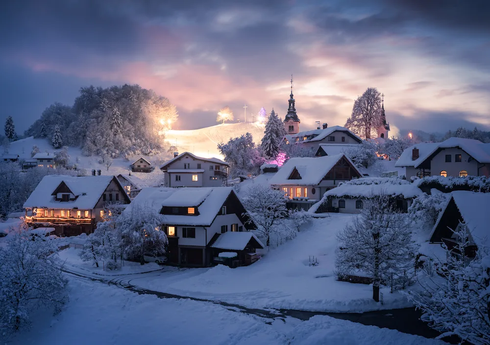  A picturesque winter village in Slovenia at dusk, featuring traditional houses with glowing windows and snow-covered roofs, a church spire in the background, and a softly lit hillside under a purple and orange sunset sky.