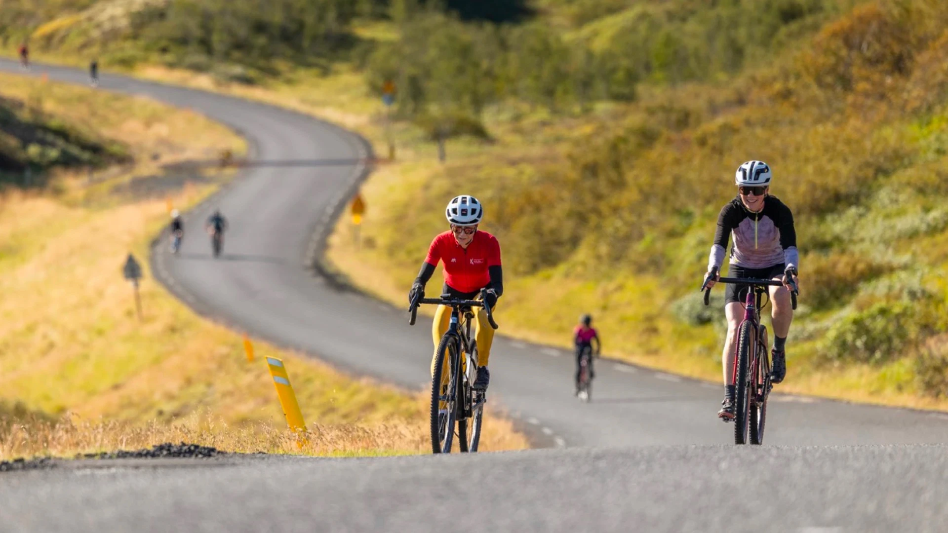two women ride their bike towards camera on a winding ashalt road