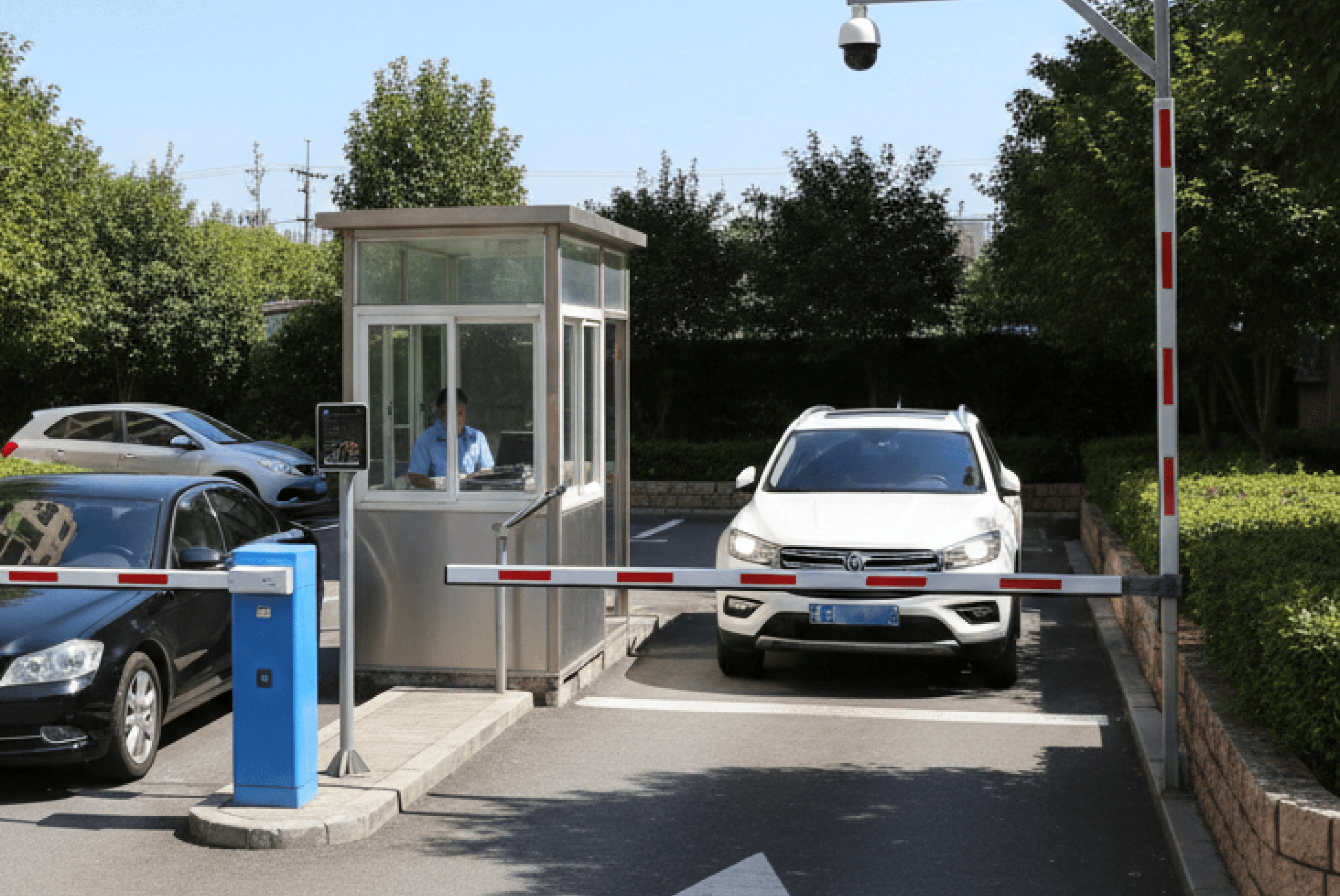 주차장 입구의 관리 초소 안에 관리 직원이 상주하고 있으며, 그 앞으로 차량 두 대가 차단기 앞에서 대기하거나 진입하는 모습으로, 파란색 주차 관리 기기와 수동 제어가 가능한 차단기가 설치되어 있습니다.