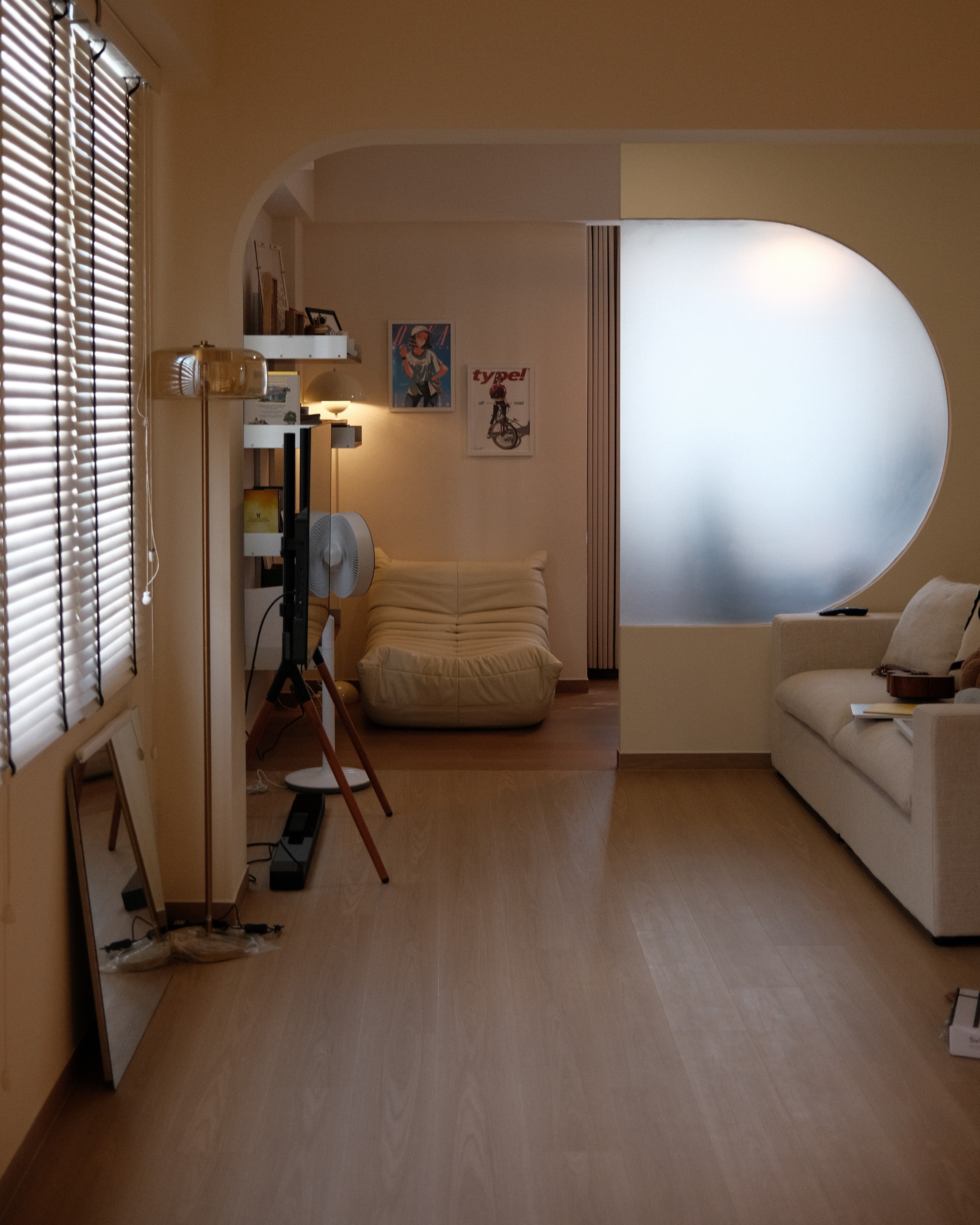 Open-concept living room at Satu-Circle with a statement circular frosted glass wall, wooden flooring, and abundant natural light from large window blinds.
