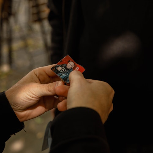 A person holding and inspecting a small photo of someone in their hands, with blurred outdoor background and another person.