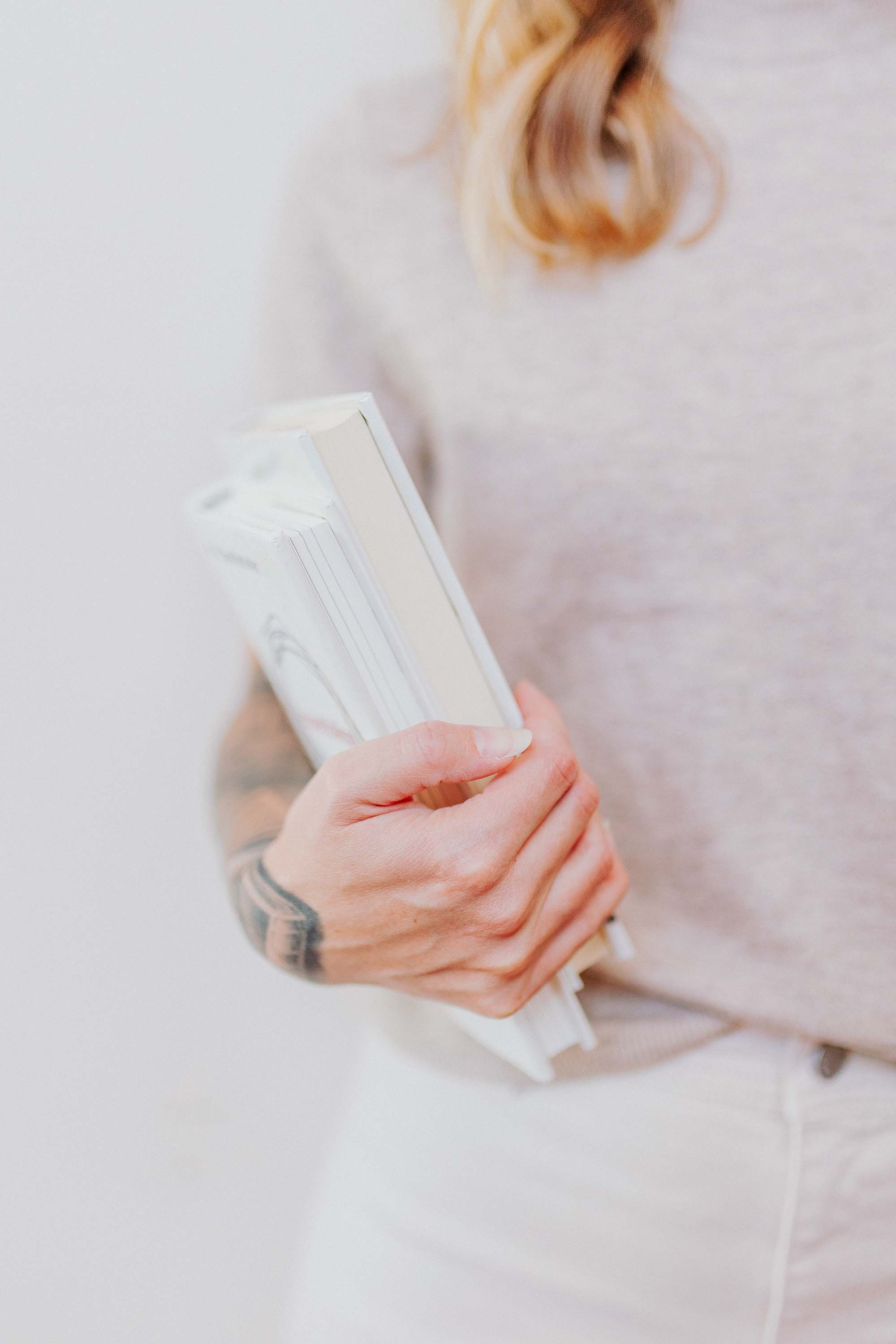 A white woman with a light grey tshirt, white jeans, and her arm covered in tattoos is holding a stack of white books in her arms
