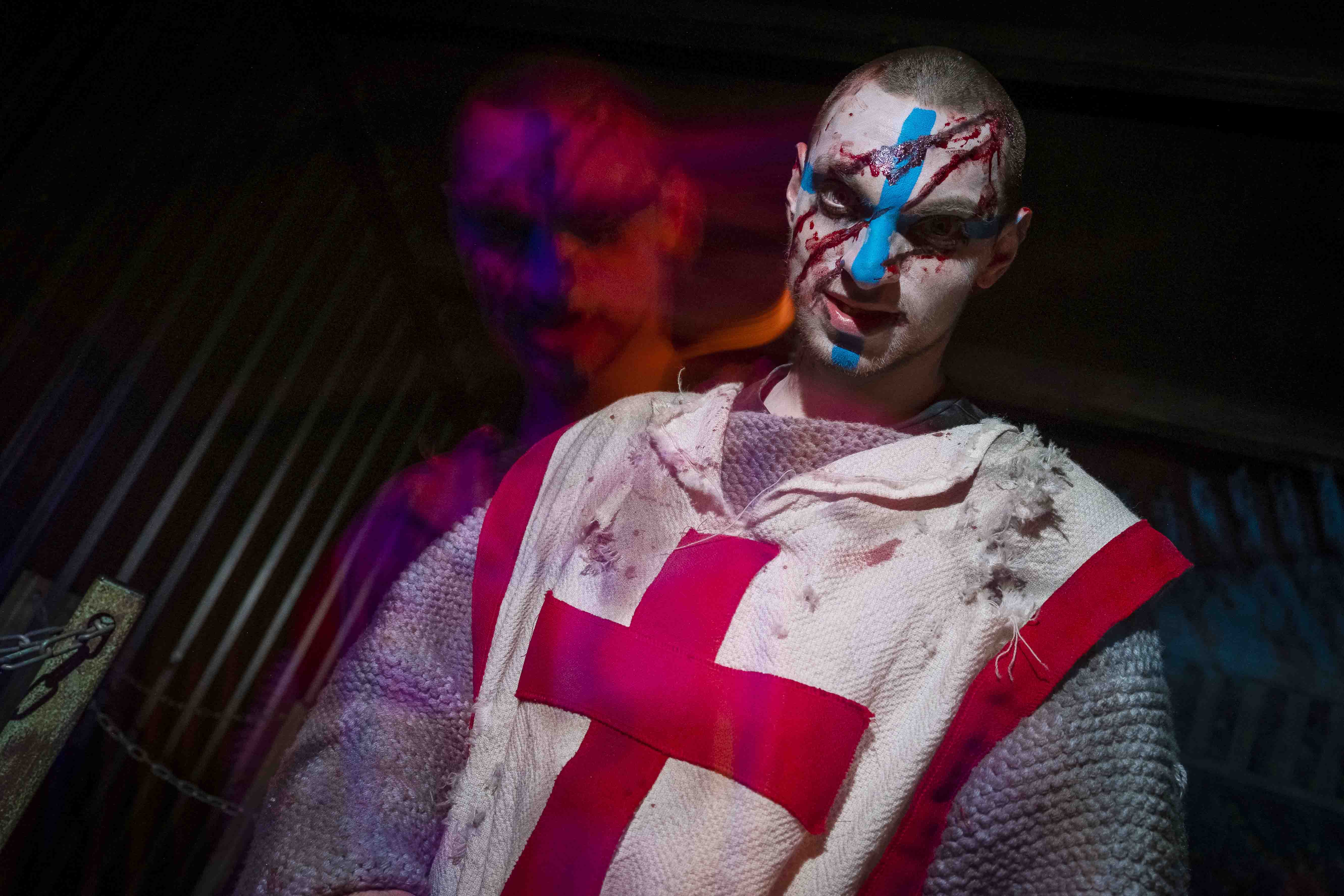 A person in a tattered, medieval-style knight costume with a red cross, painted face in red and blue, standing in a dimly lit, atmospheric scene with colorful lighting effects enhancing the dramatic and eerie setting.
