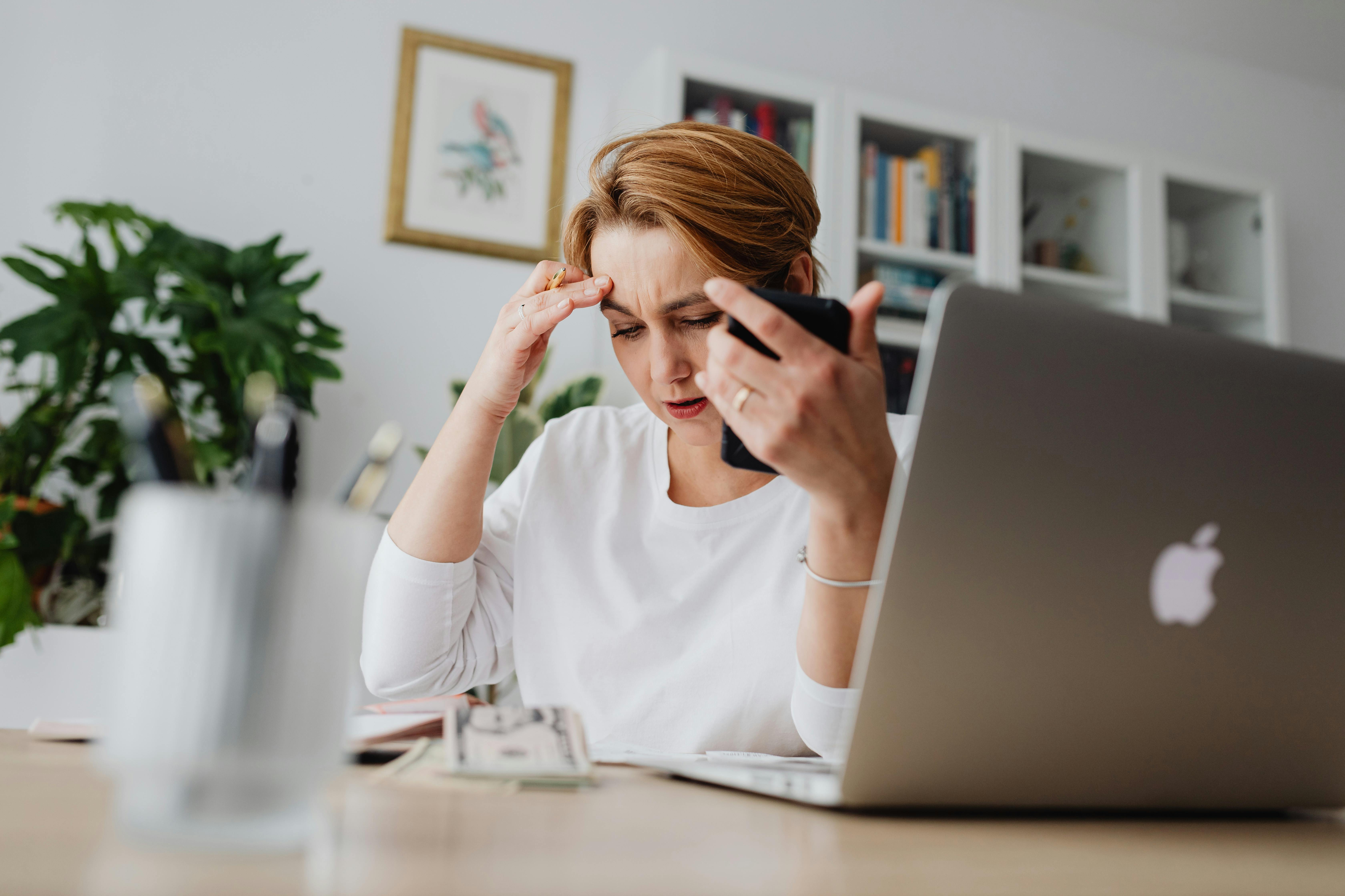 A stressed woman sitting at a desk with a laptop and phone, looking at cash and bills, illustrating the anxiety of avoiding rental scams and managing move-in costs in Canada.