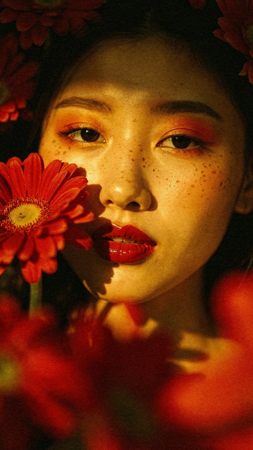 A woman with long dark hair and bold red lipstick looks over her shoulder, framed by vibrant red flowers, creating a warm and intimate mood.