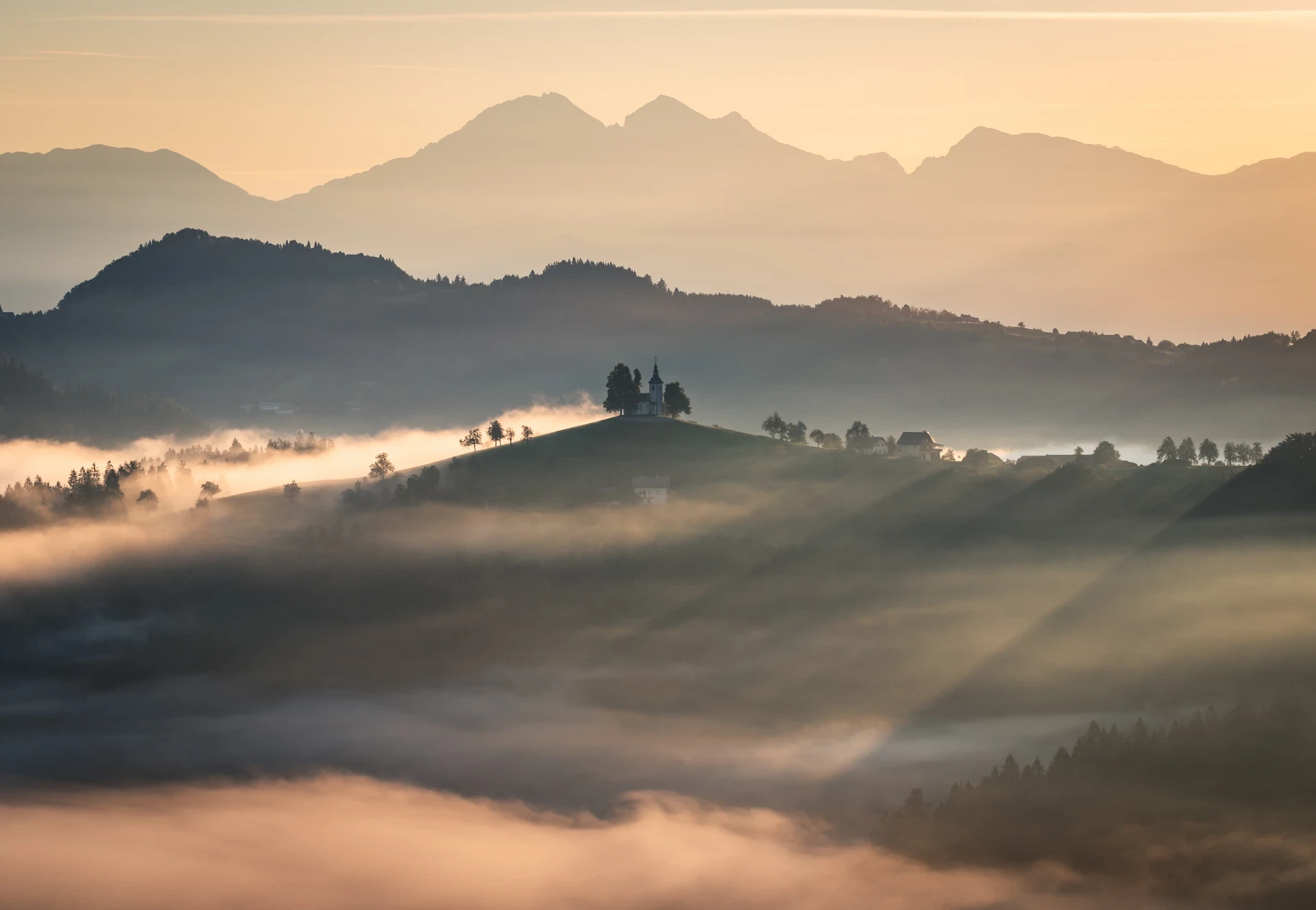 A serene landscape of Sveti Tomaž, Slovenia, featuring a lone church on a misty ridge with warm morning light filtering through a layer of low-lying clouds.