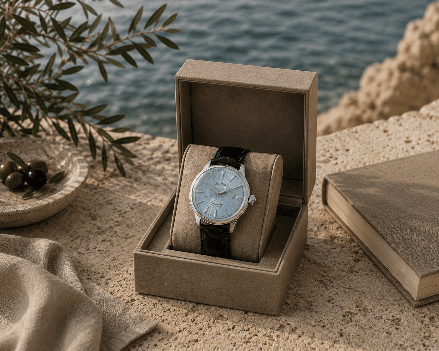 A Seiko Presage Cocktail Time watch featuring an ice blue sunburst dial, silver hour markers, and a dark blue leather strap displayed inside an open taupe suede presentation box. The watch box sits on a travertine balcony ledge overlooking blue ocean waters and rocky cliffs, styled alongside a hardcover book, a small stone dish containing olives, olive branches, and draped beige linen under natural sunlight.