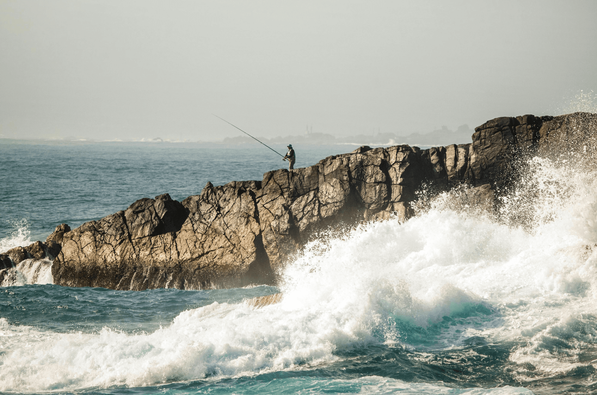 A rocky shore with waves crashing against it, a person stands on the rocks gazing at the ocean.