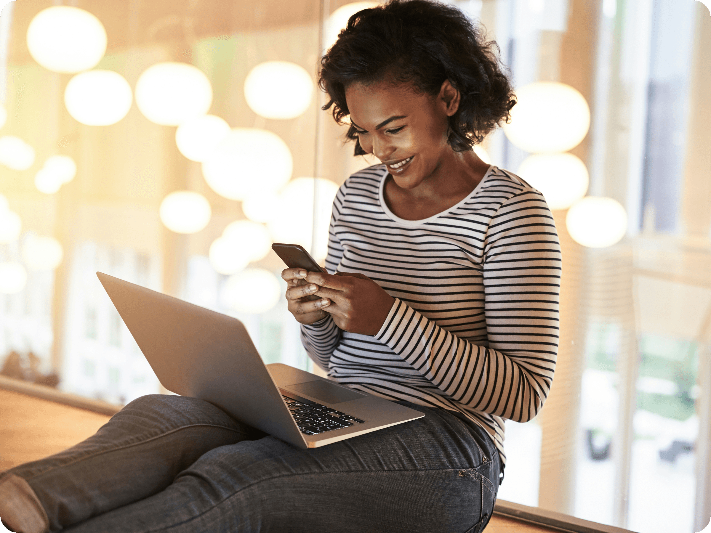 Photograph of a woman sitting on the floor of a well-lit hallway with a laptop on her lap, looking at her phone and smiling