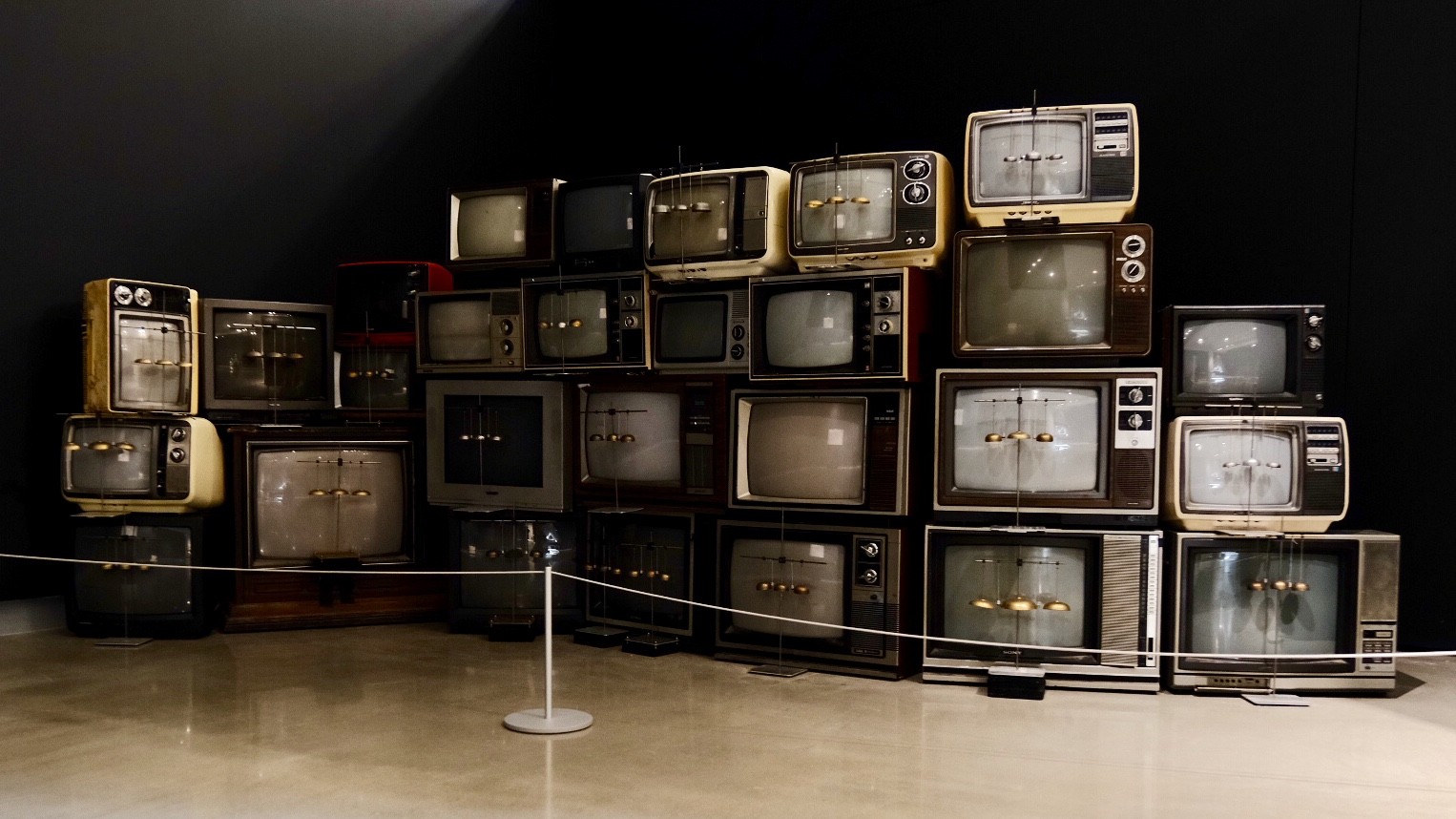 A museum installation titled 'Electrostatic Bell Choir' featuring a large wall of stacked vintage cathode-ray tube televisions from the mid-20th century. Each TV screen displays a small suspended pendulum or bell mechanism, suggesting the sets generate electrostatic fields to animate the bells. The televisions vary in size, color, and style — ranging from cream and yellow to dark wood-toned models — and are arranged in a roughly pyramidal formation against a dark wall. A rope barrier in the foreground indicates this is a gallery exhibit.
