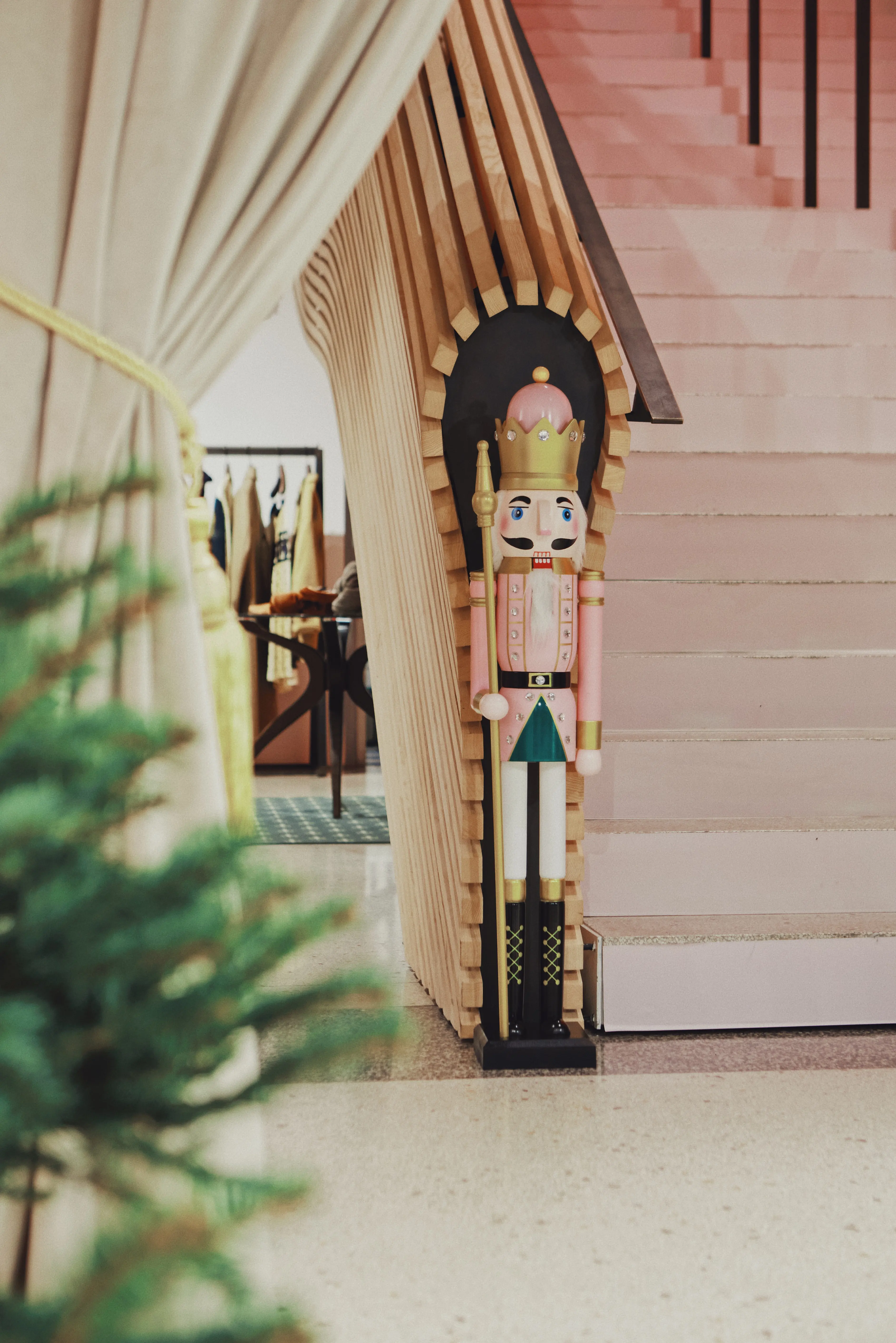 Pink-and-gold nutcracker figure standing in an arched niche of layered wooden slats beside the blush-pink staircase at UZWEI, framed by a cream curtain.