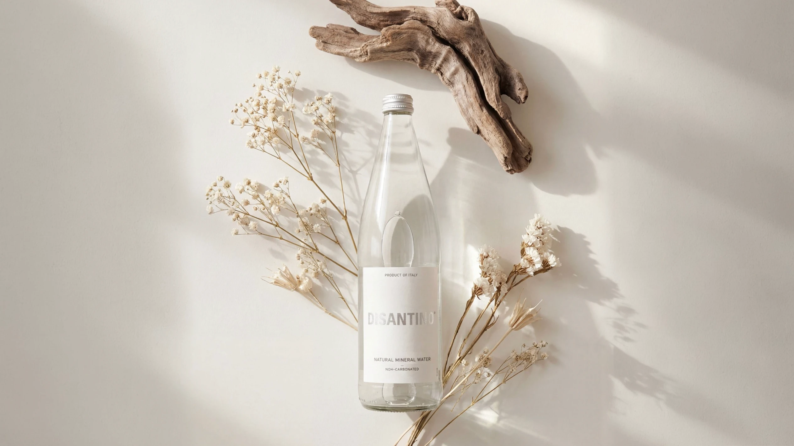 Top-down lifestyle shot of a 750ml glass bottle of Disantino Non-Carbonated water lying on a cream-colored surface. The bottle is surrounded by organic textures, including sprigs of delicate white dried flowers (baby's breath) and a weathered piece of natural driftwood. Soft, dappled sunlight and shadows fall across the scene, creating a clean and tranquil aesthetic.