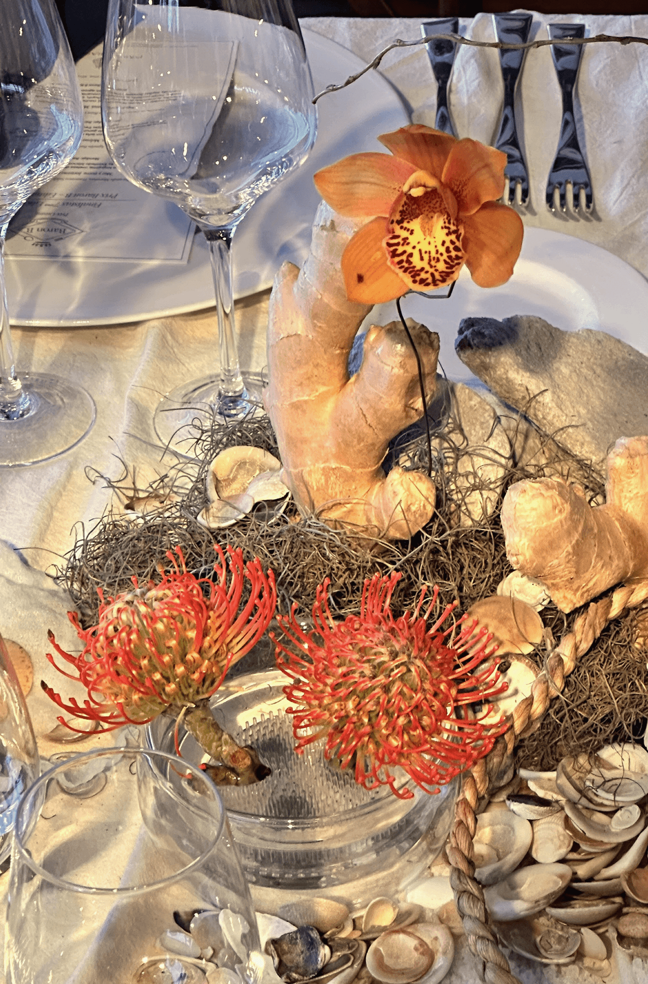 Centro de mesa con raíces de jengibre, orquídea naranja, proteas rojas y conchas marinas sobre lino crudo.