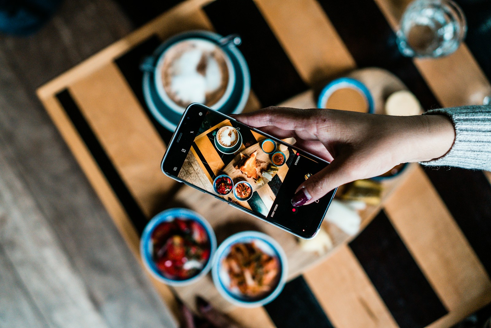 Mão segurando celular fotografando uma tábua de comida em mesa de restaurante.