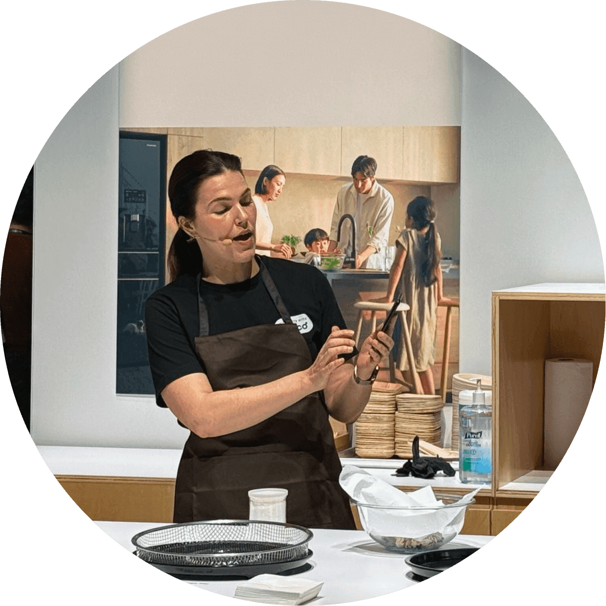 Woman in an apron demonstrates a cooking technique while using a smartphone during a Fresco product demo, with a screen behind showing people cooking together