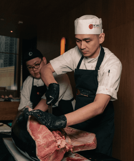 Two chefs preparing fish in a Nobu restaurant, one is skillfully cutting a large piece of tuna while another observes.
