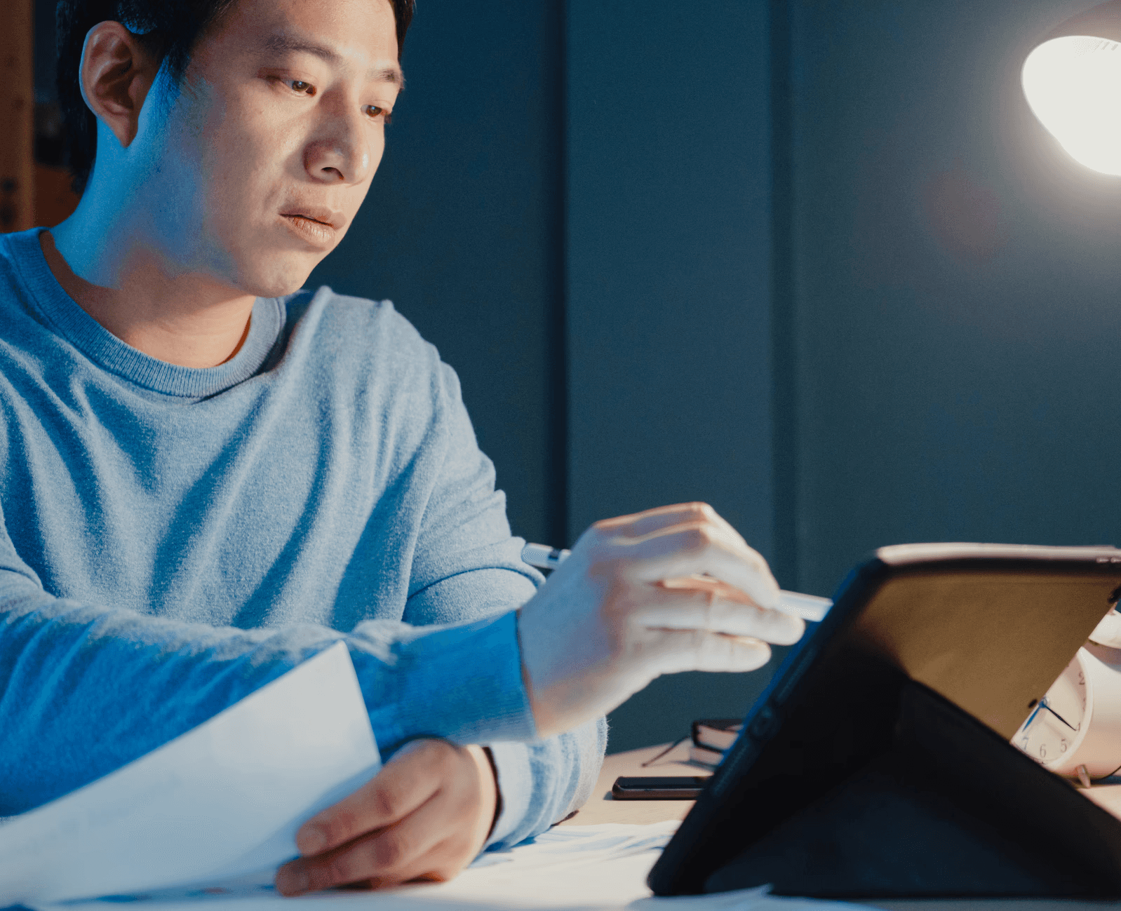 A man wearing glasses and a blazer working at a desk, writing in a notebook beside a computer monitor in a modern office.