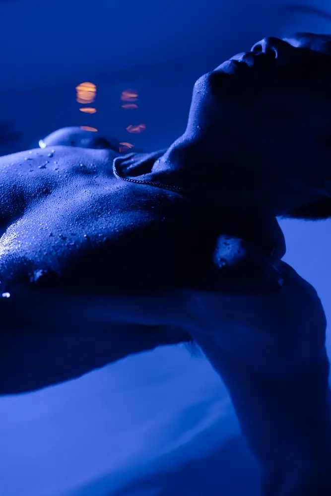 Person lying back in a cold-plunge tub with water droplets visible on the skin, lit by cool blue light.