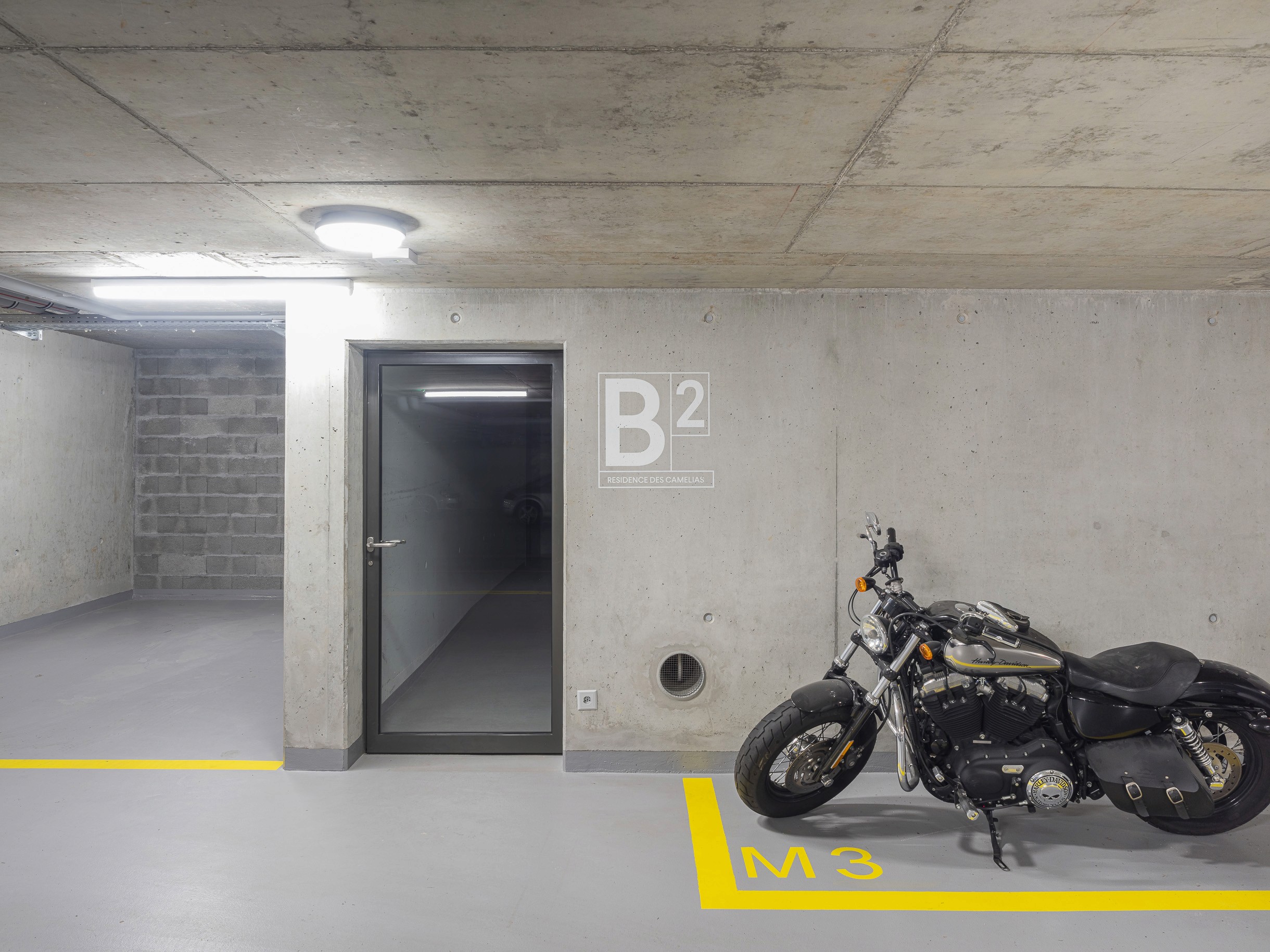 Photo d'une porte dans un garage en sous-sol et d'une moto de l amarque Harley Davidson parquée le long du mur droite sur une place de parc délimitée par un contour jaune et indiquant la place M3