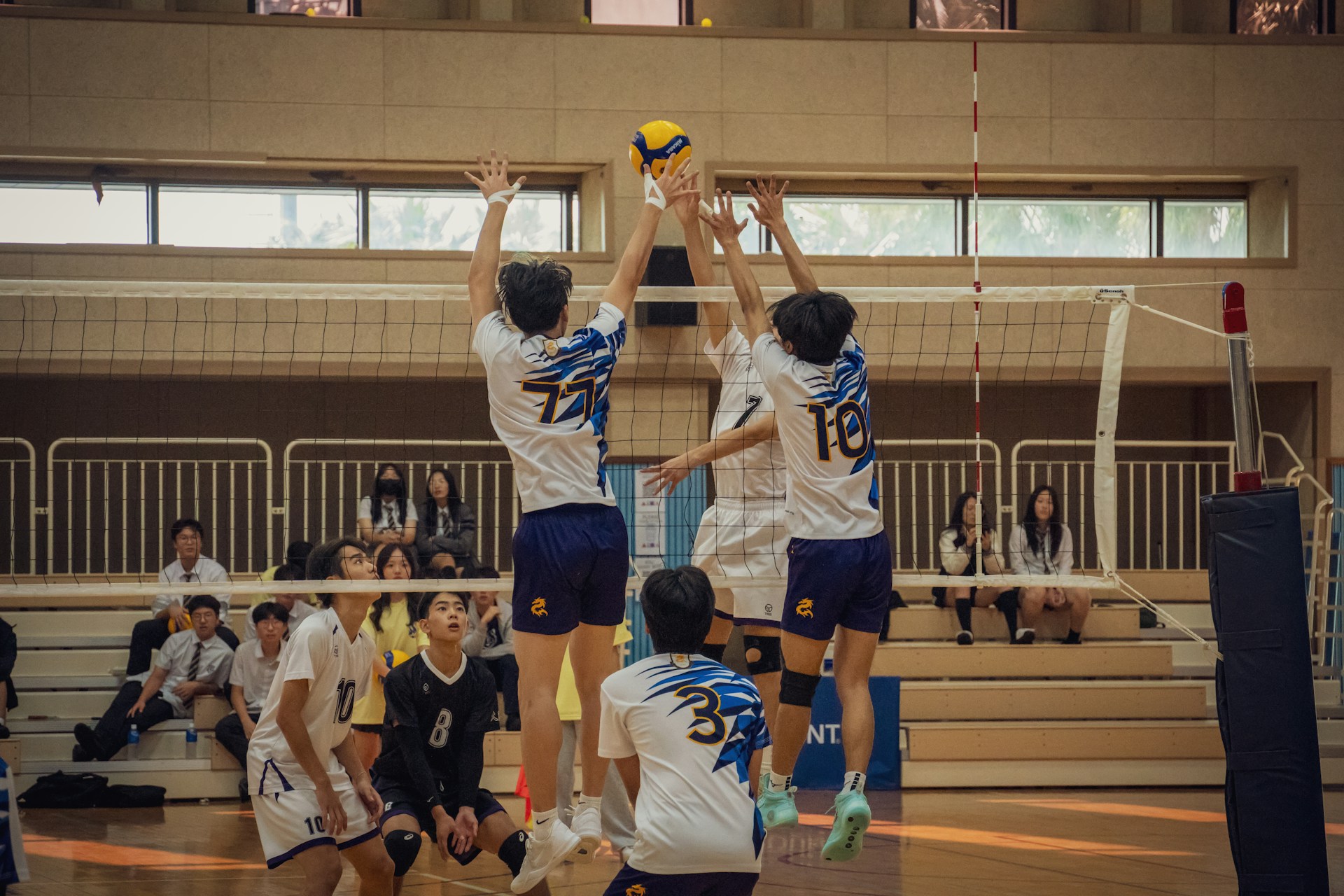 Three volleyball players jump to block a spike from an opponent during an indoor match.