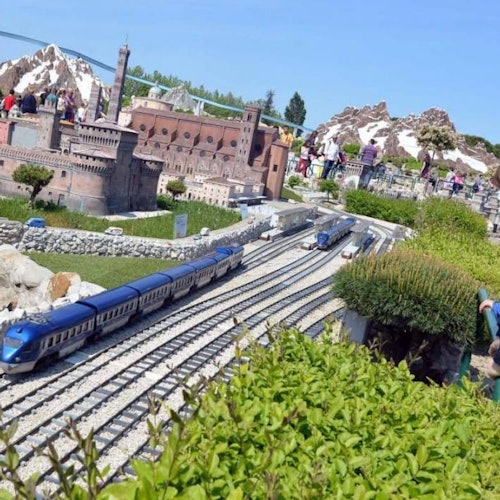People observing a large outdoor model train set with detailed buildings and landscapes under a clear blue sky.