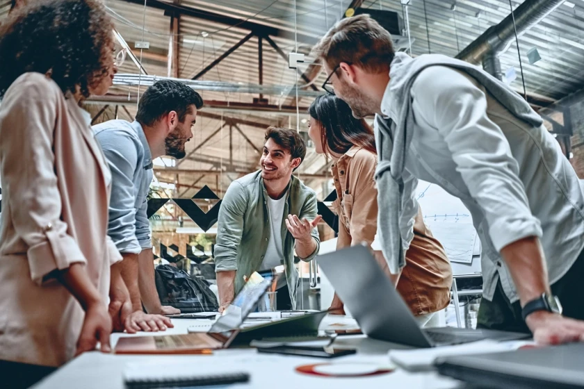 A group of colleagues engaged in a lively and collaborative discussion around a table in a modern office. The focus is on a man in the center who is gesturing as he speaks, surrounded by other colleagues who are listening attentively. The work environment appears open and creative.