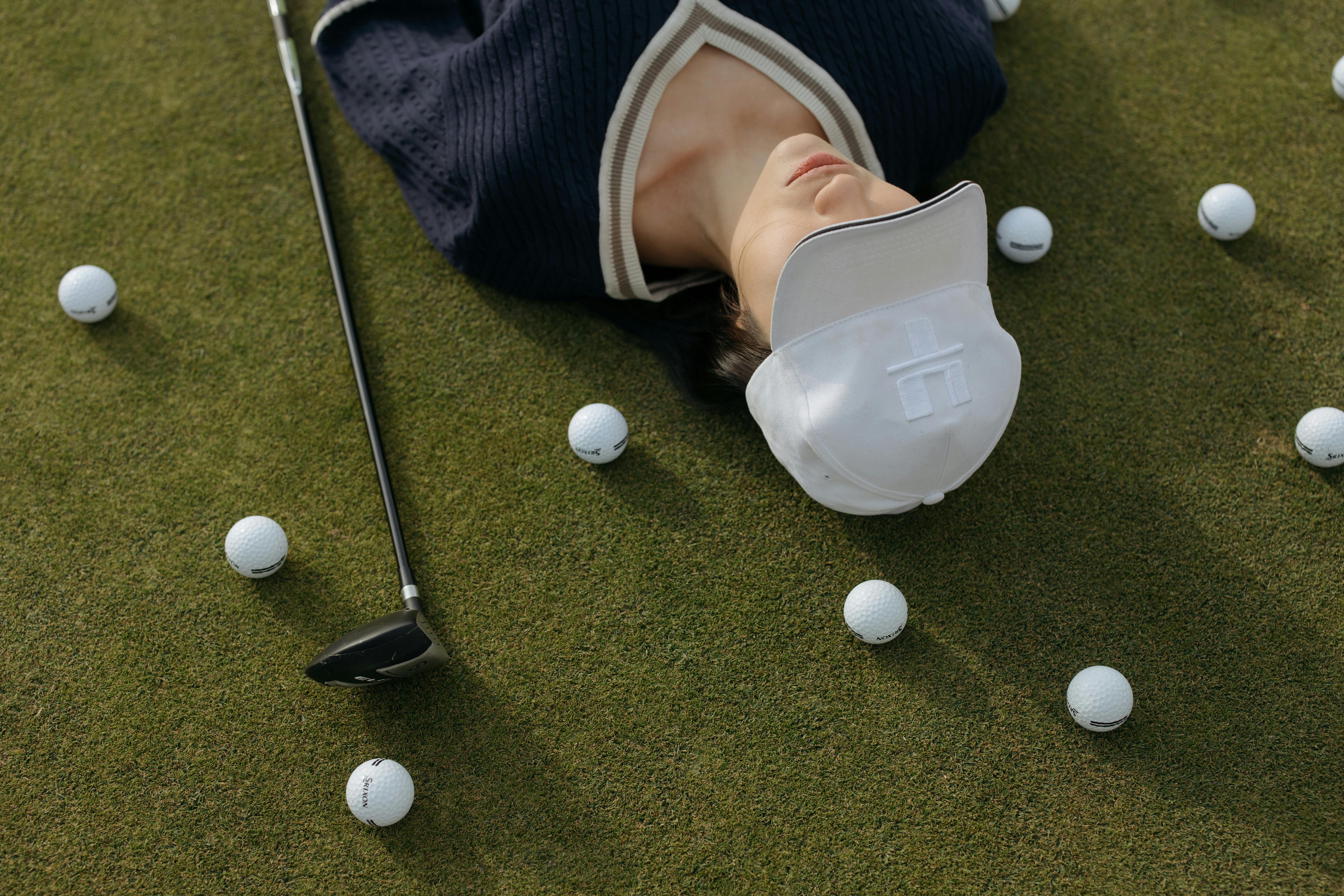 Woman lying on a golf course ground surrounded by scattered golf balls and a golf club.